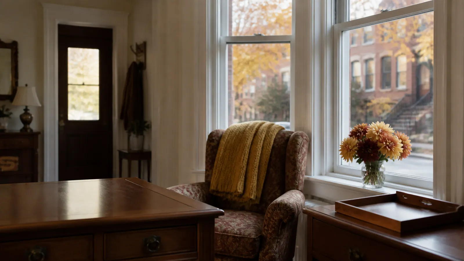 Rowhouse parlor at rest, tall windows over autumn maples, gold throw on the armchair, dahlias on the sill